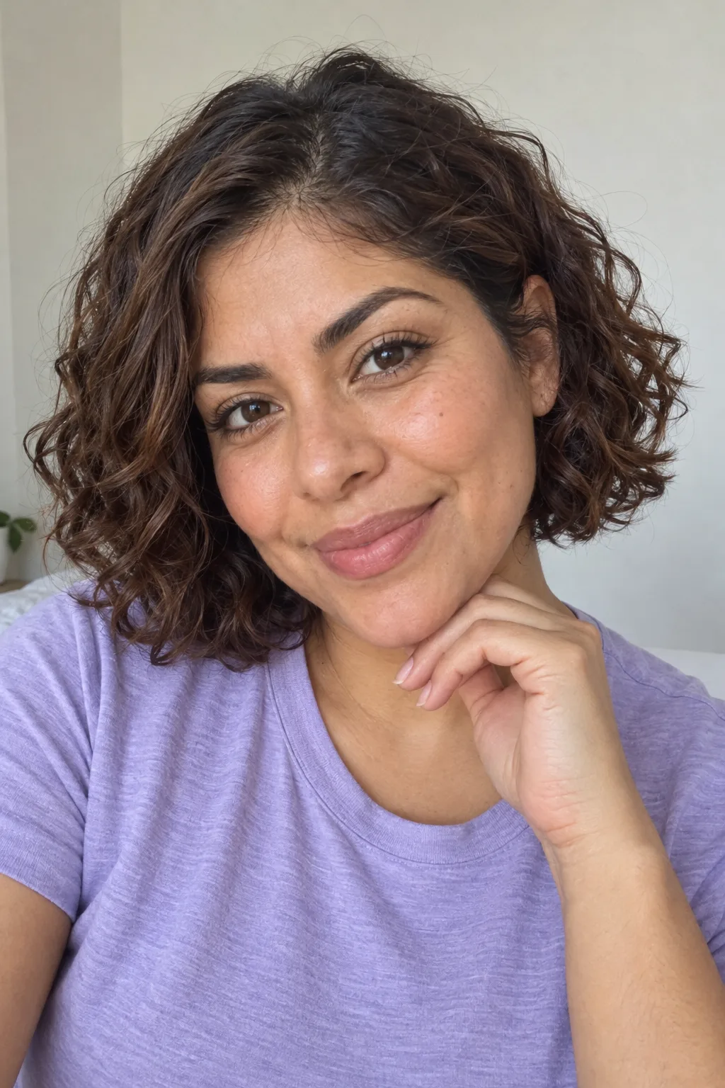 Woman with short chestnut curls smiling with her fingertips beneath her chin.