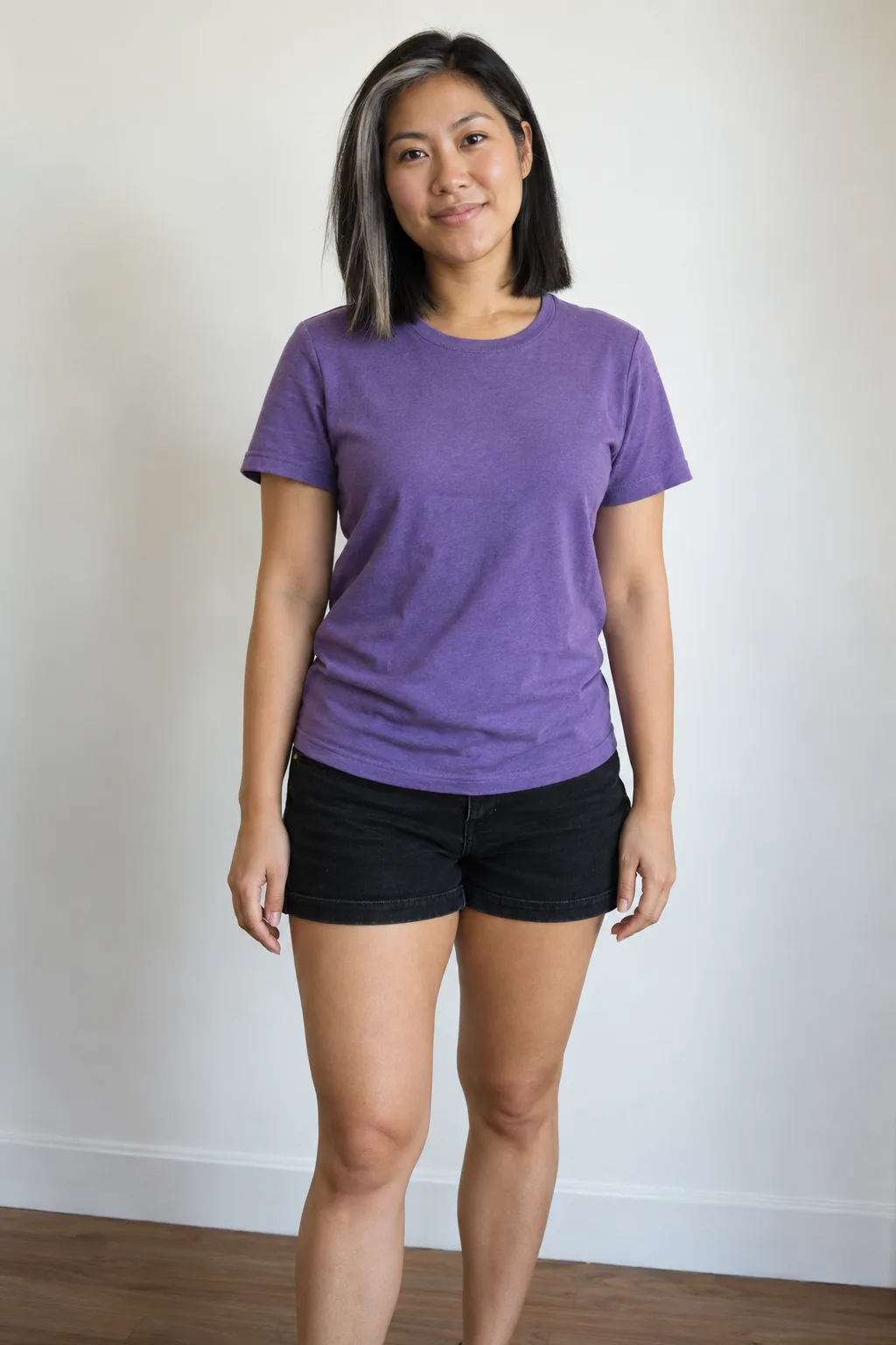 Woman with a silver-streaked dark bob standing in a violet shirt and denim shorts.